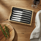Boxed steel steak knives on a wood table with rosemary, a linen towel and red wine