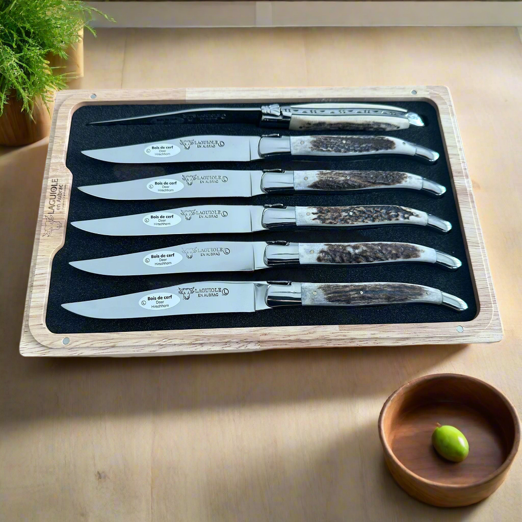 Boxed antler knives on a wooden table beside a small bowl with an olive