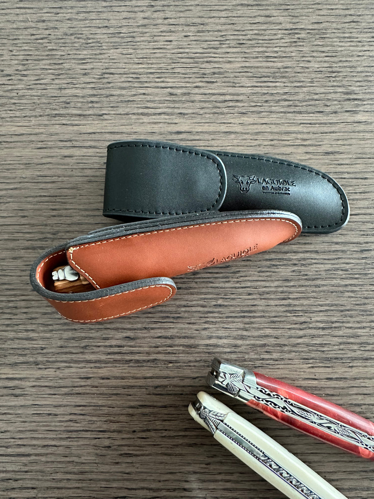 Black sheath and an open orange lined sheath beside two folding knives