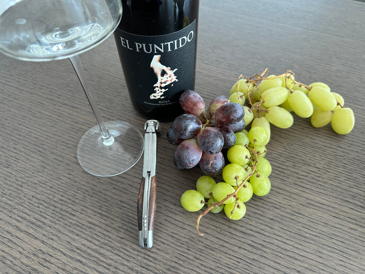 Corkscrew beside a wine glass, a bottle of red and bunches of grapes