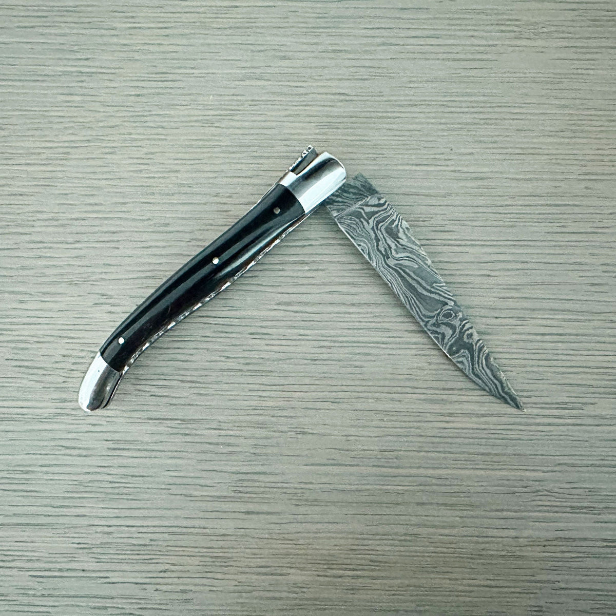 Laguiole Explosion Damascus buffalo horn knife half open on pale wood