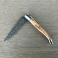 Laguiole Explosion Damascus olive wood knife half open on pale wood