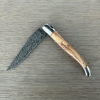Laguiole Explosion Damascus olive wood knife half open on pale wood