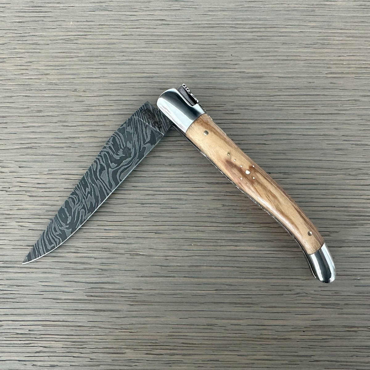 Laguiole Explosion Damascus olive wood knife half open on pale wood