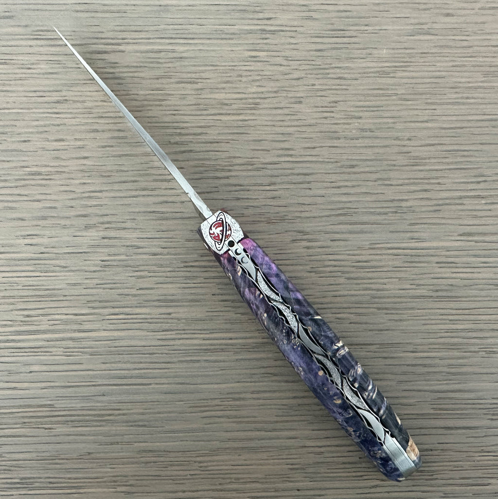 Laguiole Cosmos Saturn purple knife with indigo elder burl handle, open on pale wood