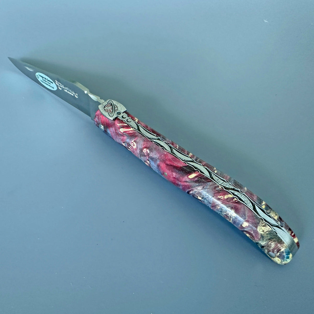 Laguiole Cosmos Saturn fire knife with red and pink elder burl handle on a blue background